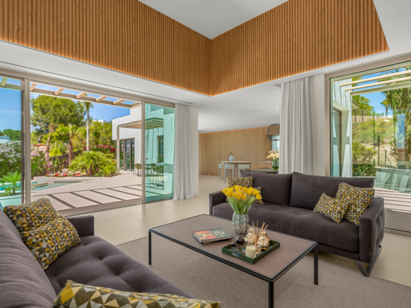 Open-plan living room with a dark brown sectional, yellow geometric cushions, and a glass door opening to a sunny backyard with a pool and lush plants outside.