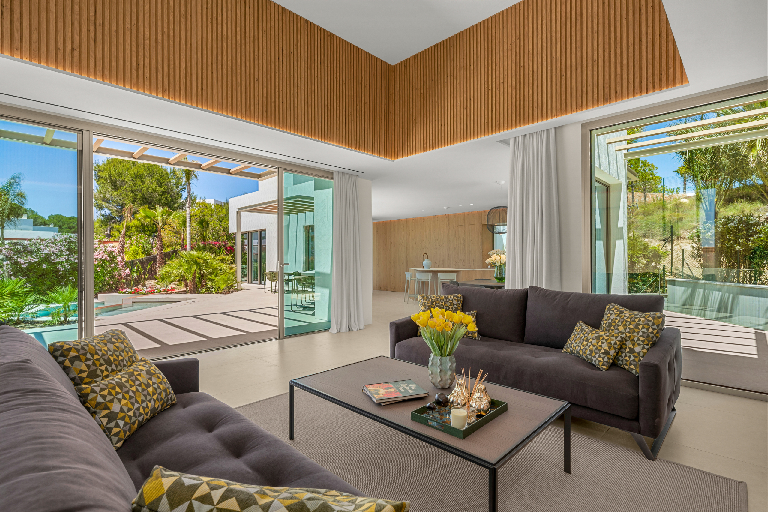 Open-plan living room with a dark brown sectional, yellow geometric cushions, and a glass door opening to a sunny backyard with a pool and lush plants outside.