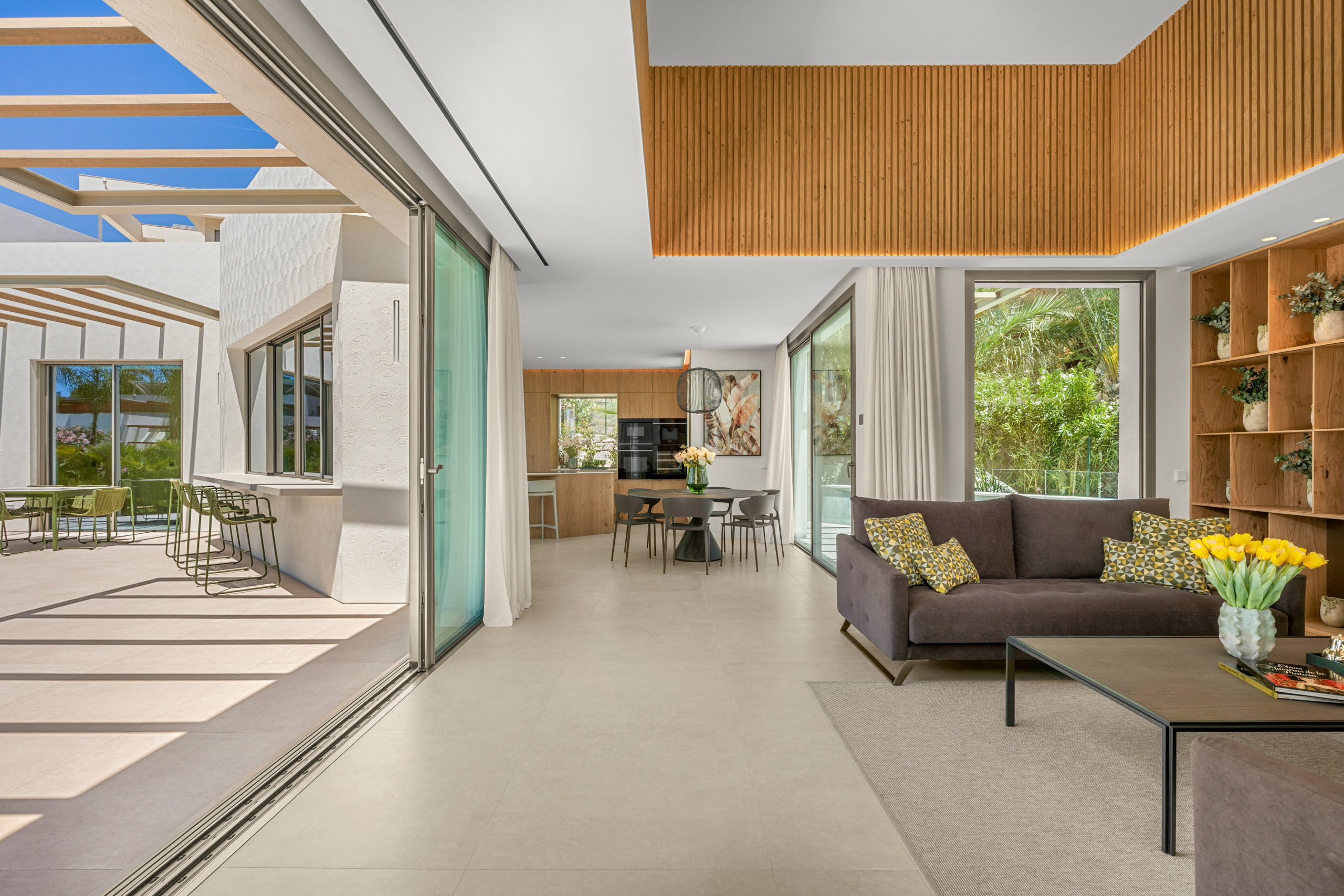Open-concept living room with a gray sofa, geometric cushions, wood shelving, and wide glass doors to an outdoor patio.