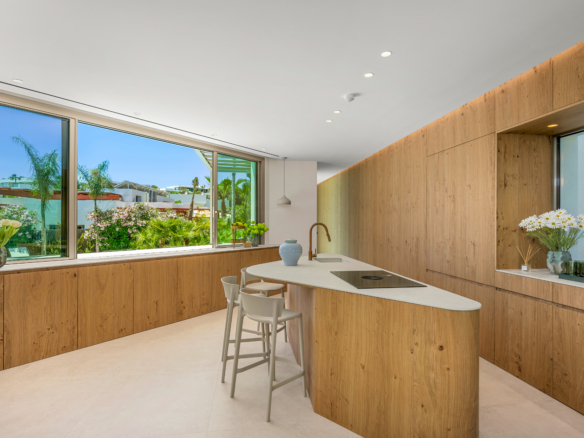 Modern kitchen with light wood cabinetry, white countertops, and a large window wall overlooking a lush garden.