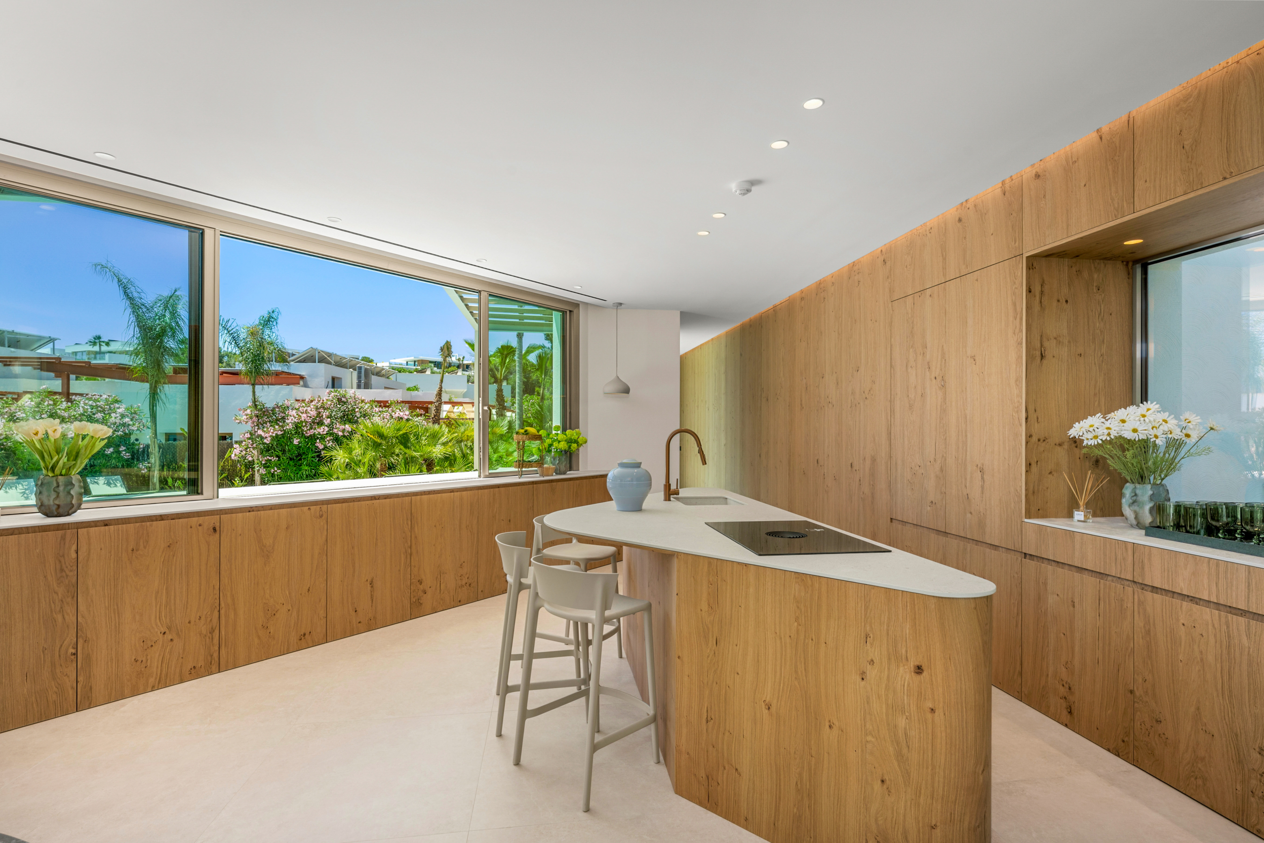 Modern kitchen with light wood cabinetry, white countertops, and a large window wall overlooking a lush garden.