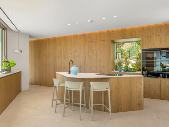 Modern kitchen with warm wood cabinets, a white island, and four white stools under an airy skylight effect; garden view through a large window.