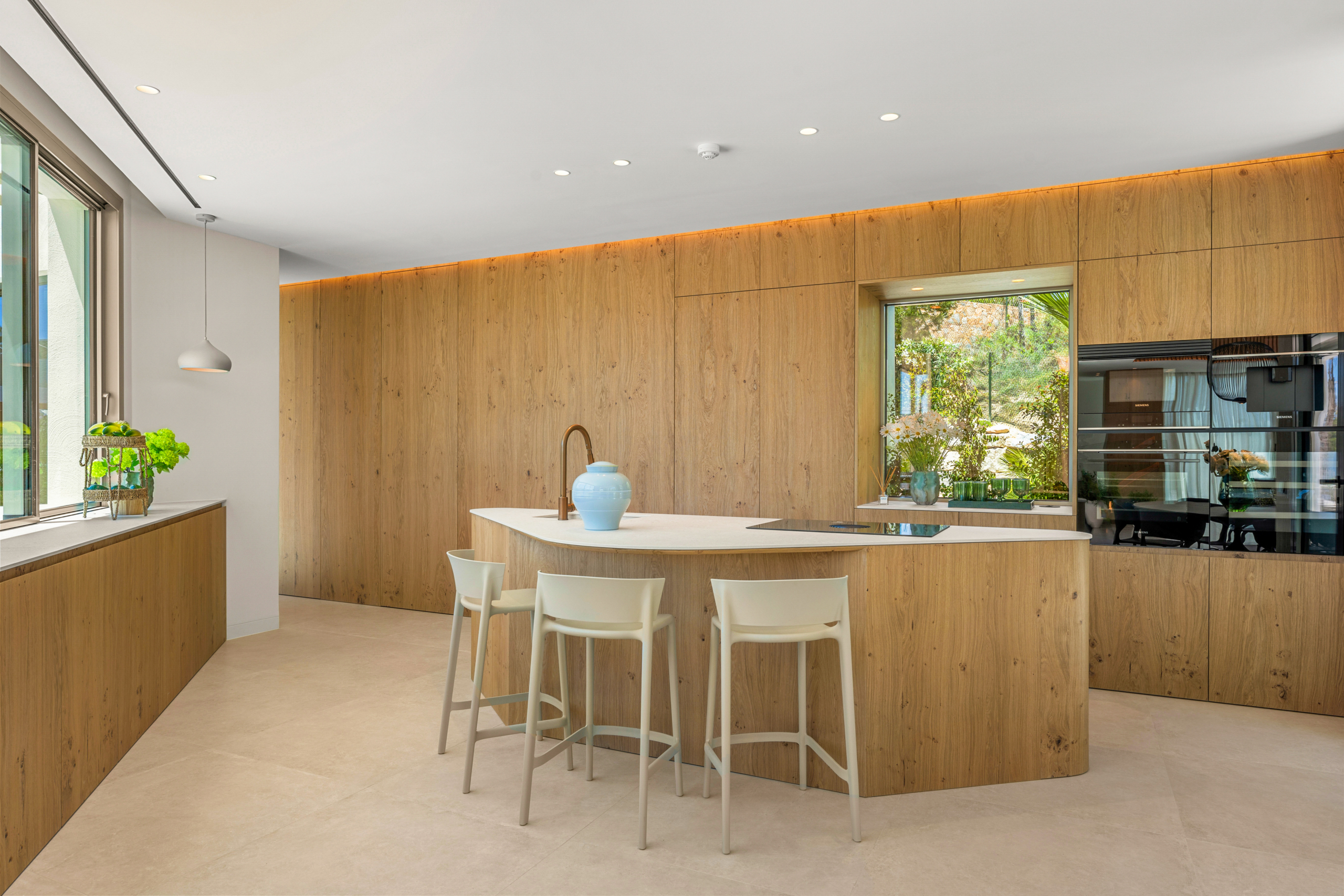 Modern kitchen with warm wood cabinets, a white island, and four white stools under an airy skylight effect; garden view through a large window.