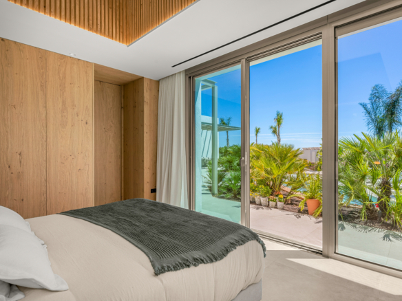 Modern bedroom with wood-paneled walls and large glass doors opening to a sunny tropical garden.