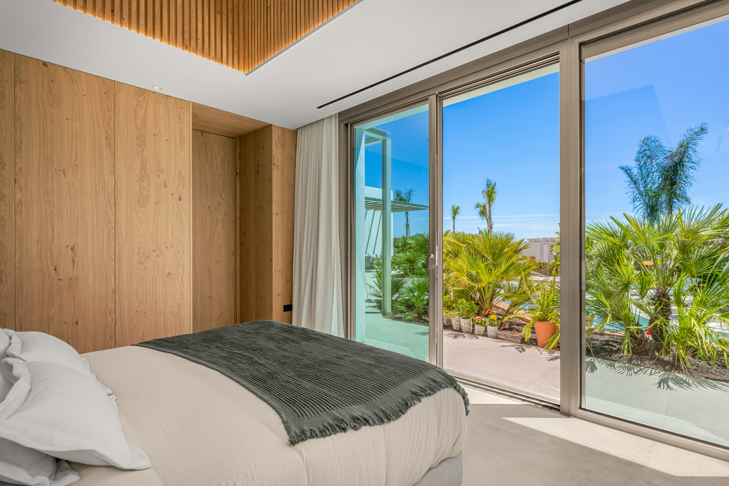 Modern bedroom with wood-paneled walls and large glass doors opening to a sunny tropical garden.