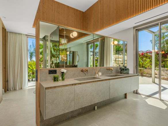 Modern bathroom with a double-sink vanity, large mirrors, and wood-accented ceiling; sliding glass doors open to a sunlit garden.