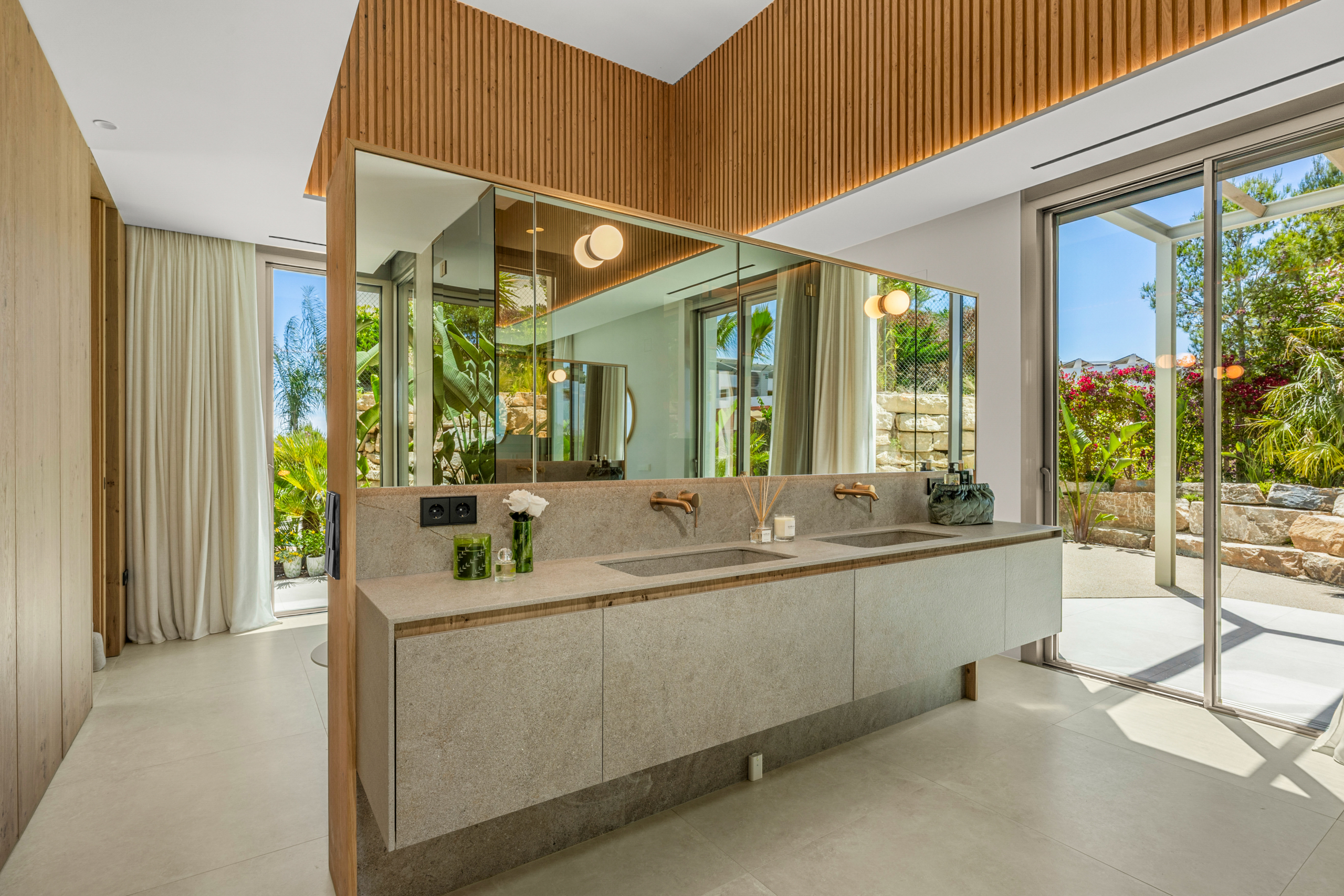 Modern bathroom with a double-sink vanity, large mirrors, and wood-accented ceiling; sliding glass doors open to a sunlit garden.