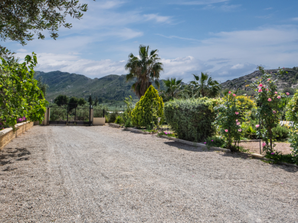 Gravel driveway leading to a black metal gate, surrounded by lush greenery and distant mountains under a blue sky