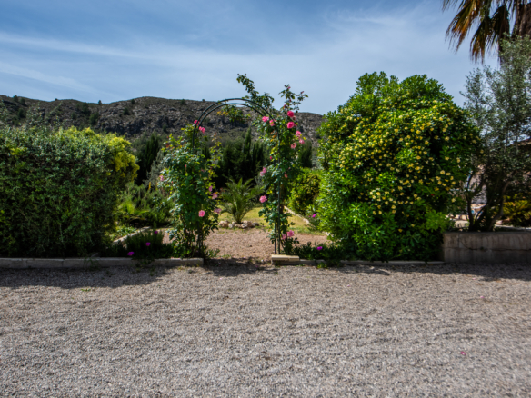 Garden arch with pink roses, green shrubbery, and a gravel foreground against a blue sky and distant hillside