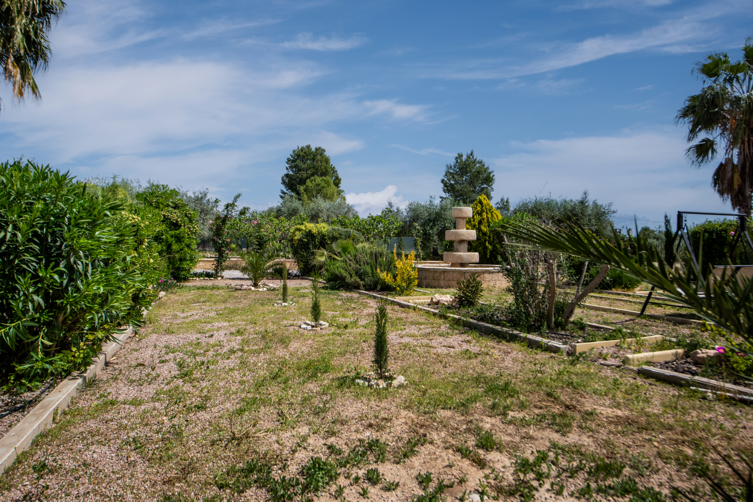 Sunny garden scene with gravel paths, lush shrubs, and a tiered stone fountain under a blue sky.