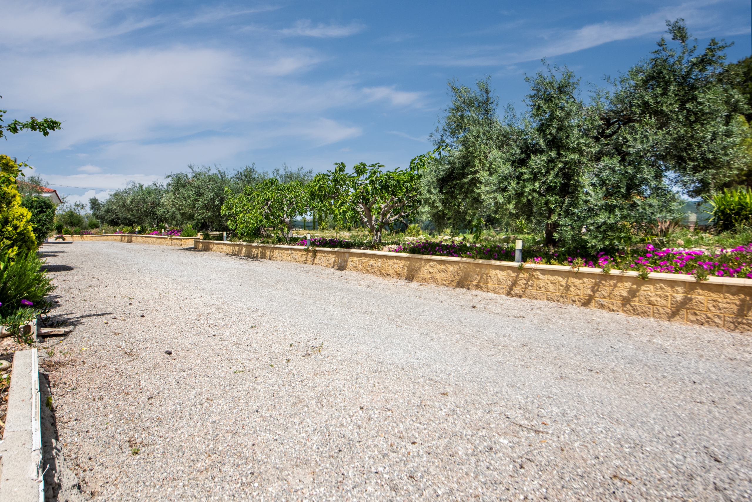 Gravel lot or garden pathway with a low stone wall, trees, and colorful flowers under a blue sky.