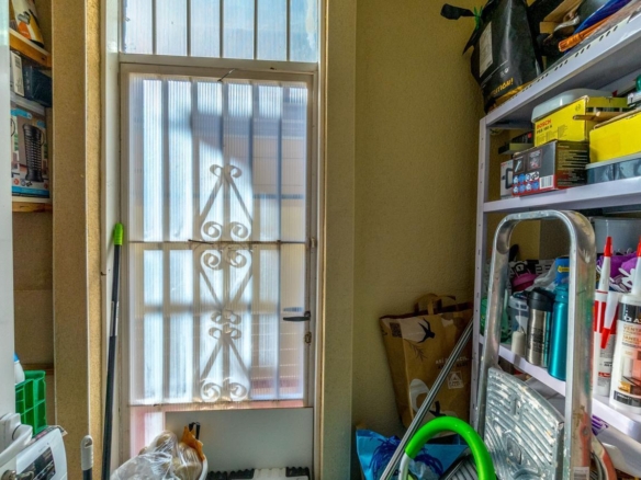 A narrow storage room with a frosted glass door and decorative metal bars, and shelves filled with boxes, bottles, and tools on the right side.