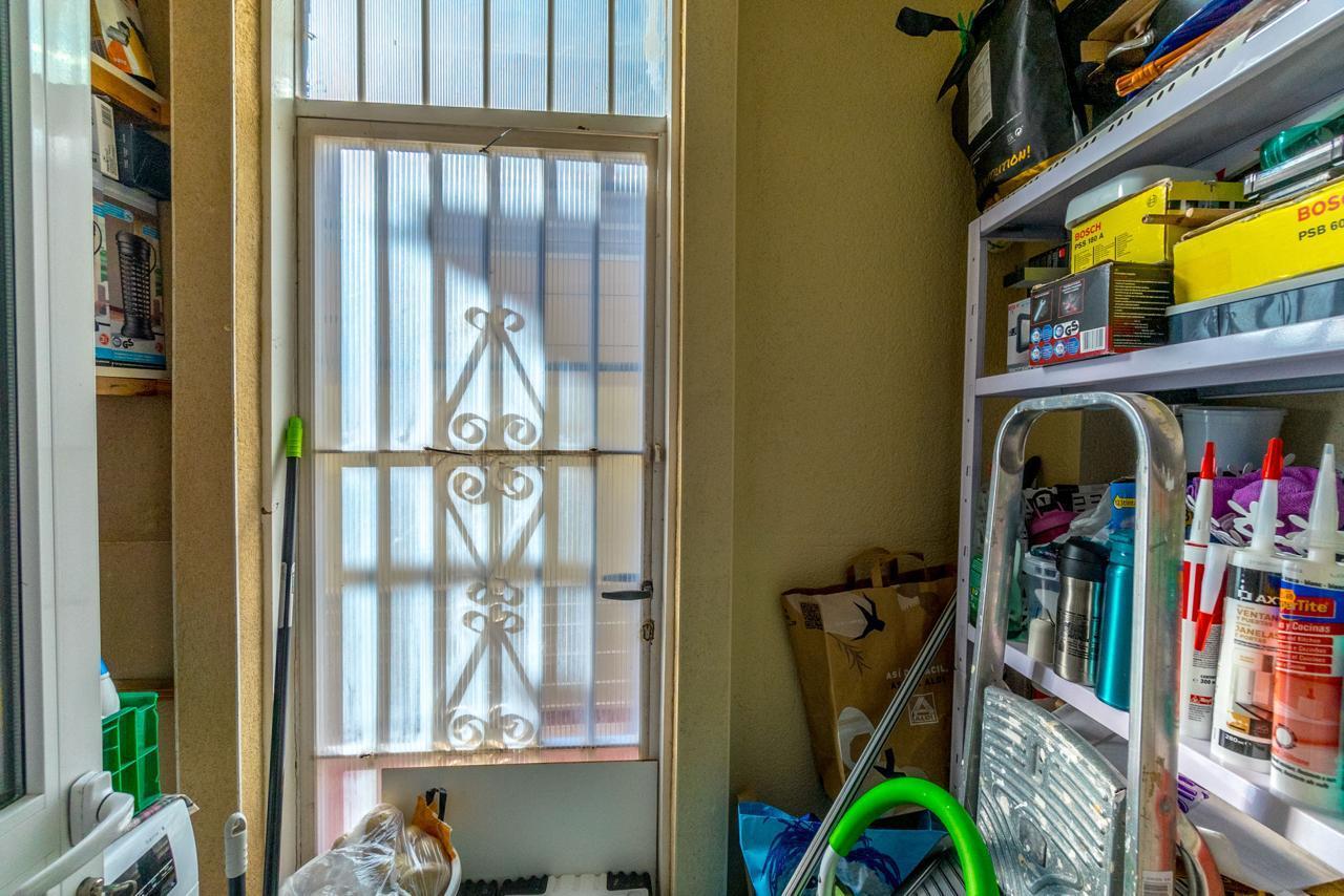 A narrow storage room with a frosted glass door and decorative metal bars, and shelves filled with boxes, bottles, and tools on the right side.