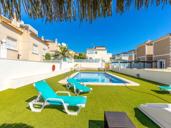 Poolside area with turquoise lounge chairs on green artificial grass, a rectangular blue pool, and beige residential buildings under a clear blue sky.
