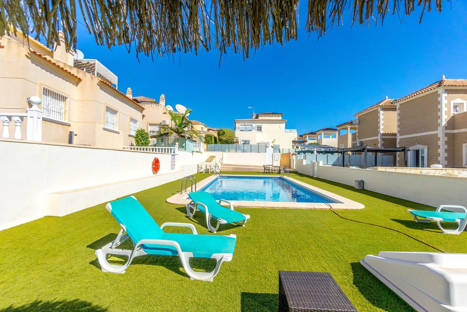 Poolside area with turquoise lounge chairs on green artificial grass, a rectangular blue pool, and beige residential buildings under a clear blue sky.