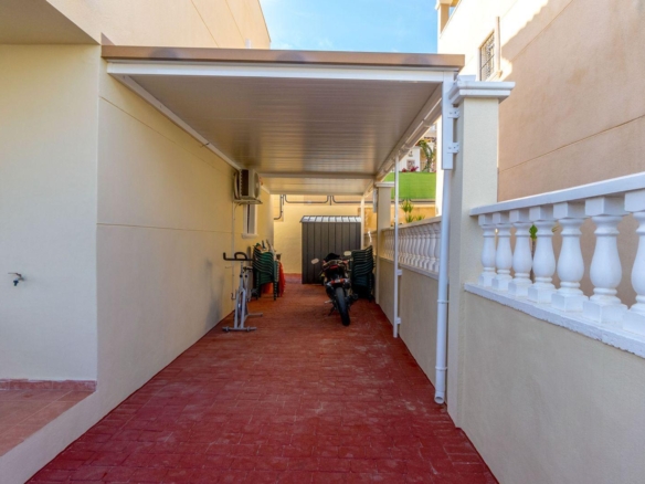 Covered outdoor corridor with red brick floor, bikes and a storage shed at the far end; plastic chairs stacked along the wall.