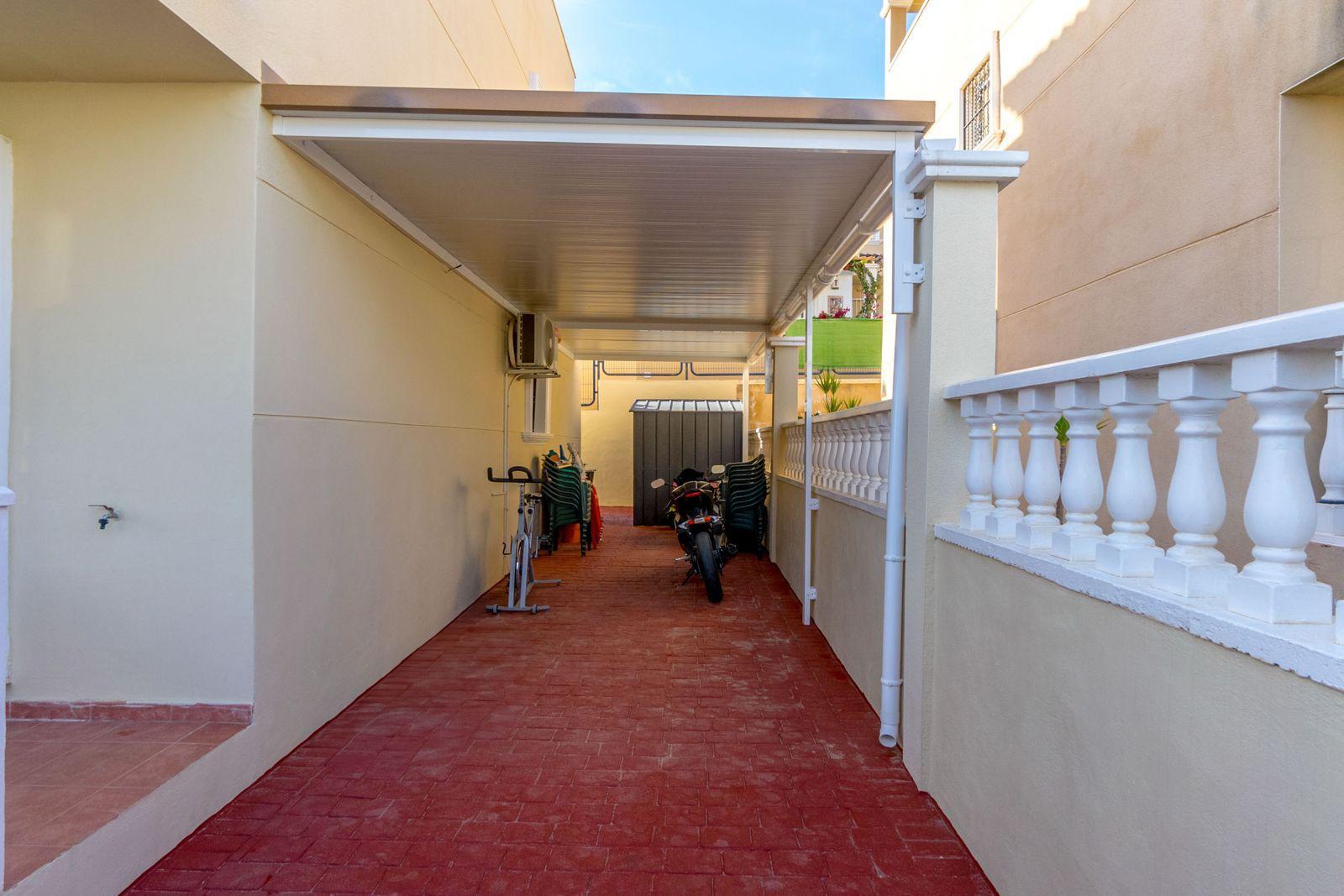 Covered outdoor corridor with red brick floor, bikes and a storage shed at the far end; plastic chairs stacked along the wall.
