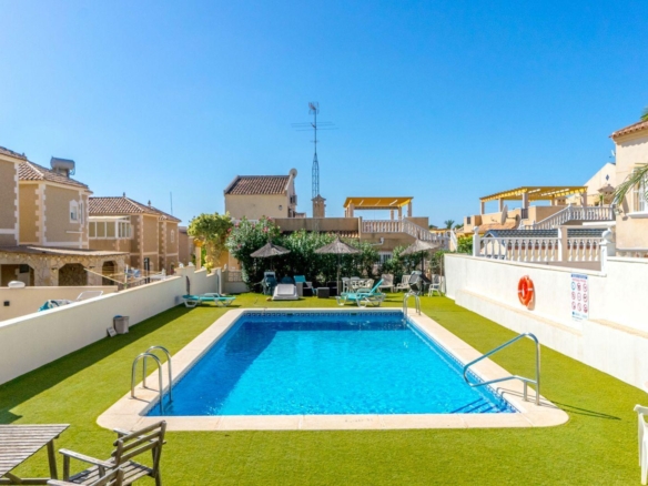 Rectangular outdoor pool at a sunny resort, surrounded by lounge chairs, thatched umbrellas, and beige buildings under a clear blue sky.
