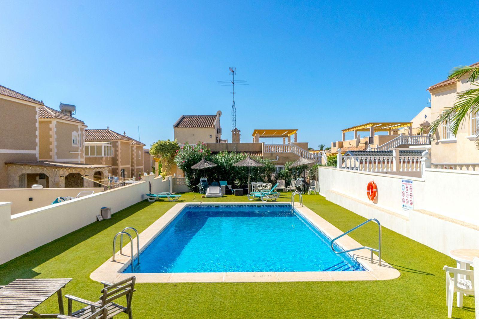 Rectangular outdoor pool at a sunny resort, surrounded by lounge chairs, thatched umbrellas, and beige buildings under a clear blue sky.