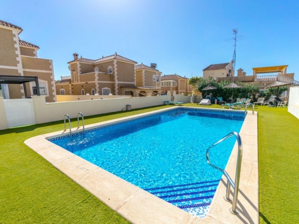 Pool in a sunny courtyard with tan houses, blue water, and surrounding green artificial grass; lounge chairs and umbrellas nearby.