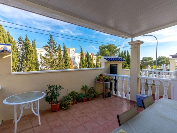 Sunlit balcony with terracotta tiles, a beige wall lined with potted plants, and a white railing. extra chairs and table nearby under a covered area.