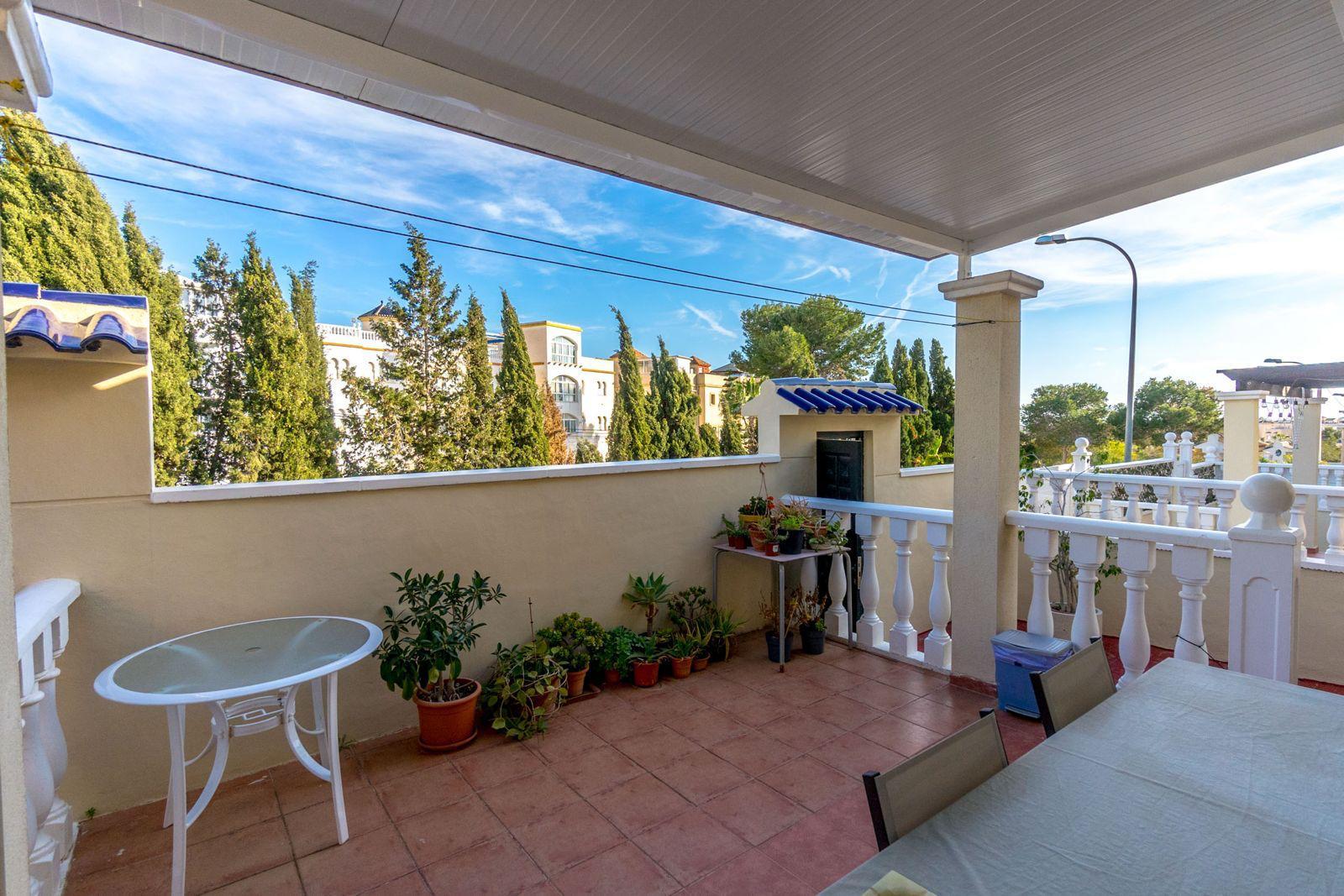 Sunlit balcony with terracotta tiles, a beige wall lined with potted plants, and a white railing. extra chairs and table nearby under a covered area.