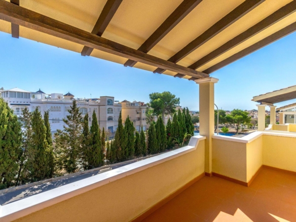 Balcony with a wooden-beam pergola, tan tiles, and warm yellow walls overlooks a row of evergreen trees and white apartment buildings against a clear blue sky.