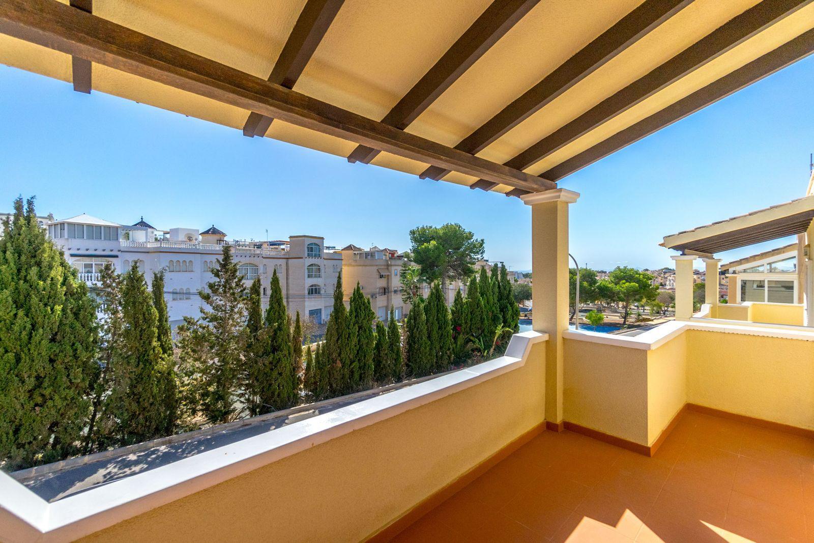 Balcony with a wooden-beam pergola, tan tiles, and warm yellow walls overlooks a row of evergreen trees and white apartment buildings against a clear blue sky.