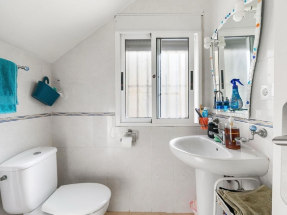 Small white bathroom with a toilet, pedestal sink, and a window above the sink; blue towel hangs on the wall.