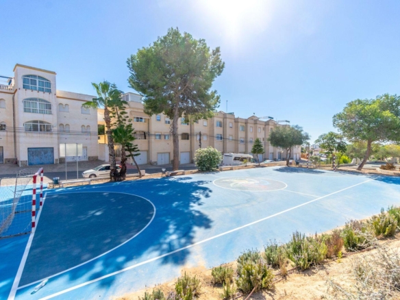 Blue outdoor sports court with white boundary lines and a small goal, set beside beige apartment buildings under a bright sky.