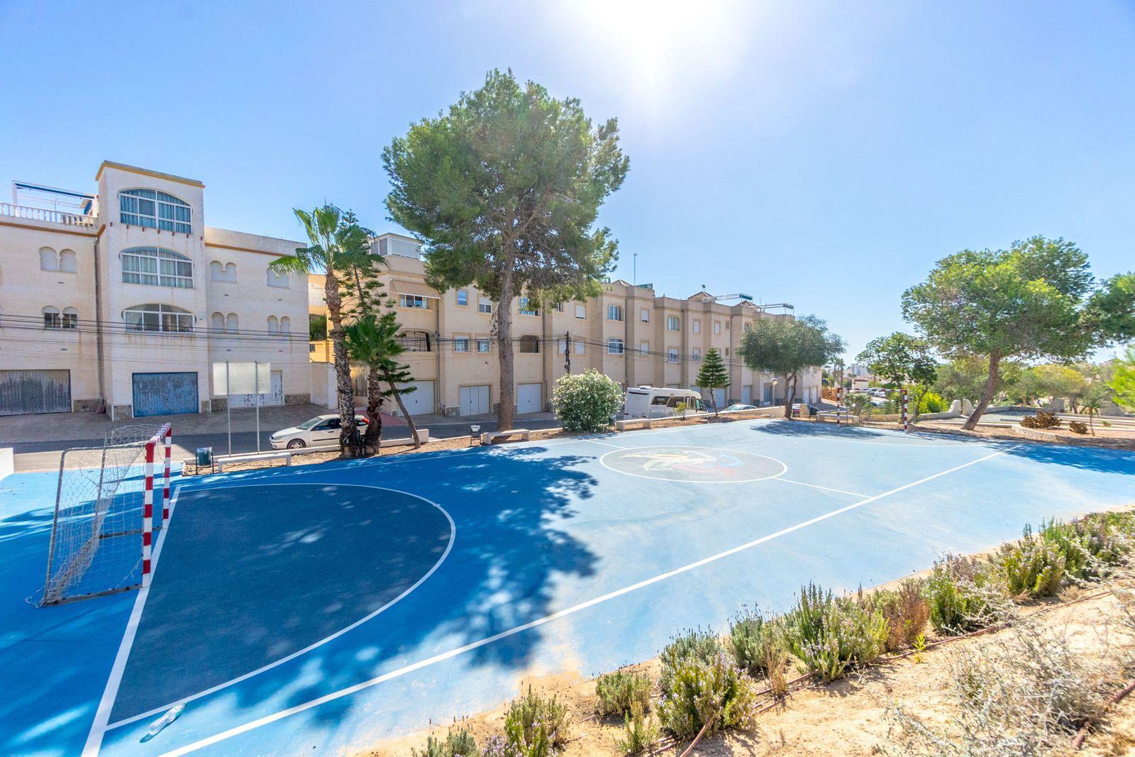 Blue outdoor sports court with white boundary lines and a small goal, set beside beige apartment buildings under a bright sky.