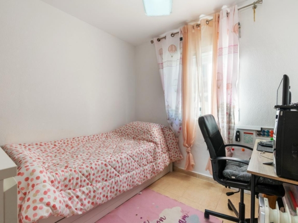Small bedroom with a single bed along the left wall, pink dotted bedding, and sheer pink curtains by the window opposite a desk and computer.