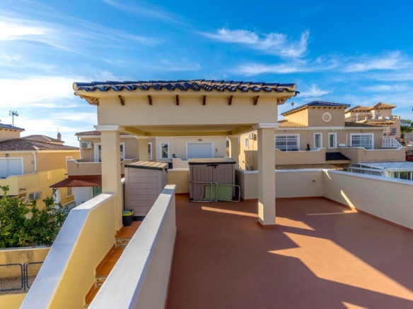 Rooftop terrace with beige columns and a blue-tiled overhang, overlooking sunlit Mediterranean-style houses under a clear blue sky.