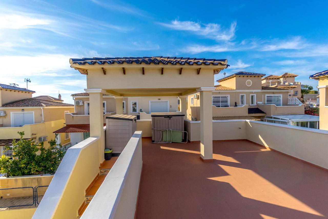 Rooftop terrace with beige columns and a blue-tiled overhang, overlooking sunlit Mediterranean-style houses under a clear blue sky.
