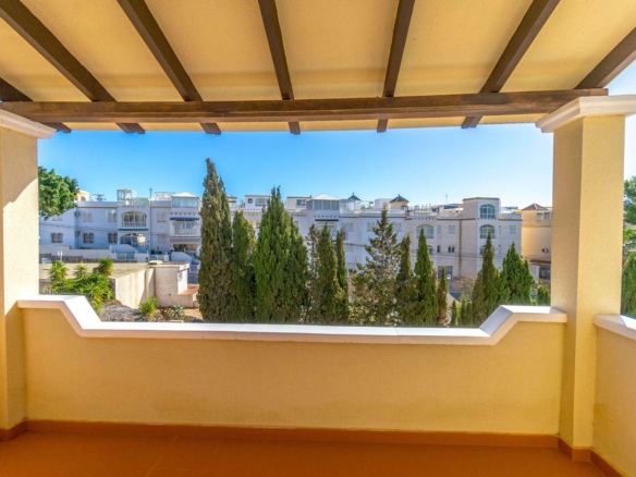 Balcony railing frames a view of white apartment buildings and tall evergreen trees under a bright blue sky.