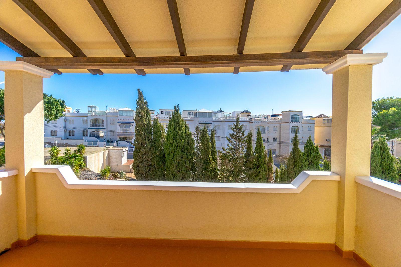 Balcony railing frames a view of white apartment buildings and tall evergreen trees under a bright blue sky.