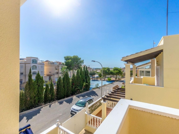 Sunny residential street view from a balcony, with beige buildings, parked car, and tall evergreen trees lining the road.