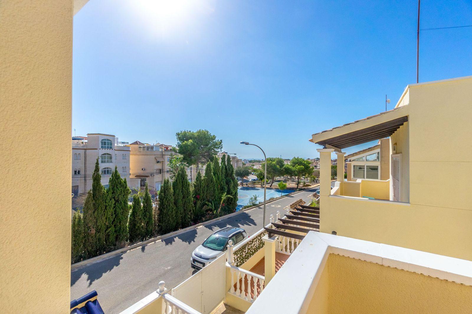 Sunny residential street view from a balcony, with beige buildings, parked car, and tall evergreen trees lining the road.