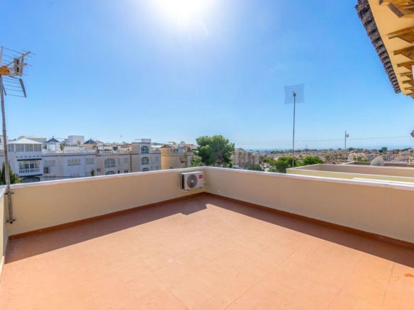 Sunlit rooftop terrace with terracotta tiles, beige railing, and an air conditioner, overlooking white apartment buildings and a blue sky.