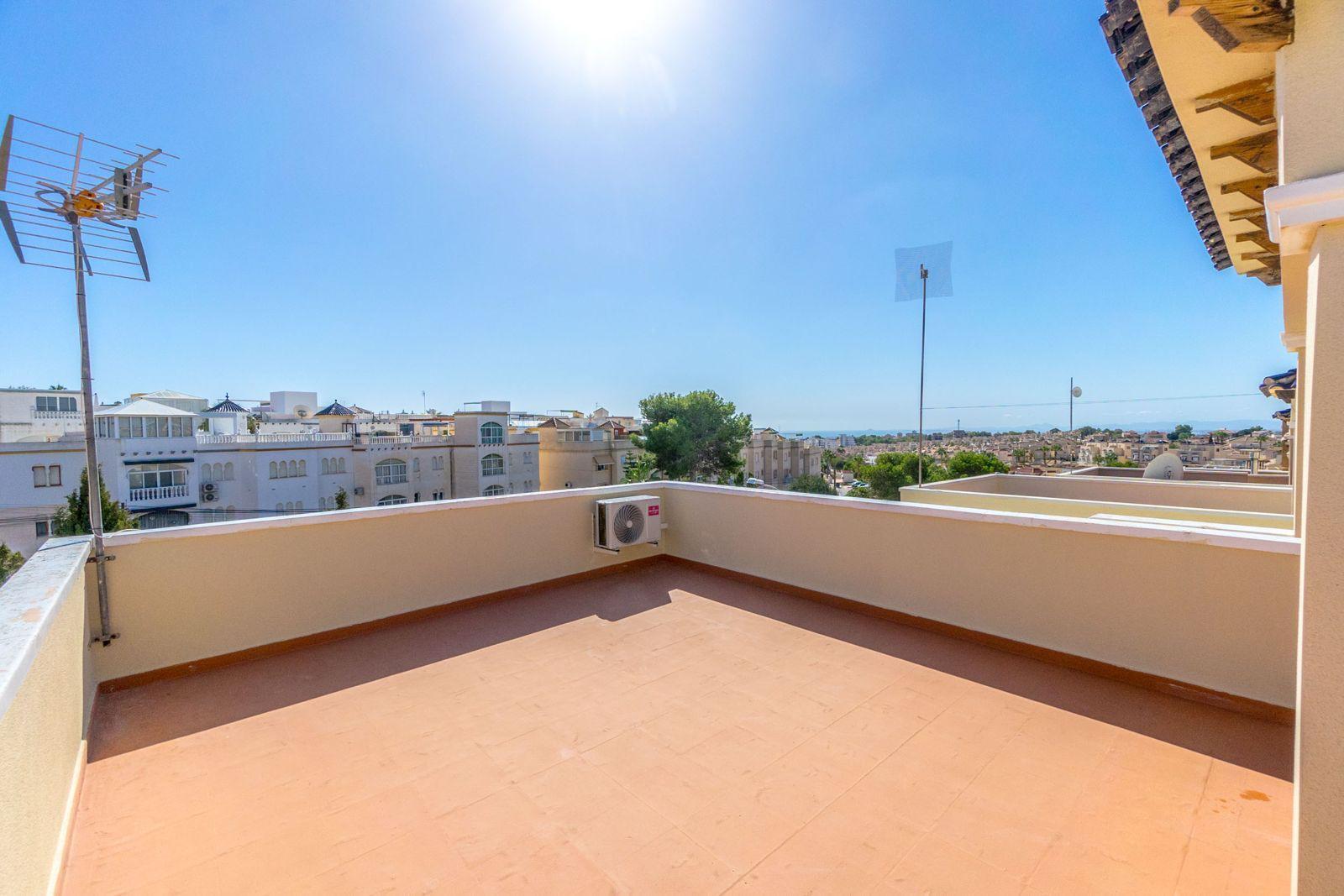 Sunlit rooftop terrace with terracotta tiles, beige railing, and an air conditioner, overlooking white apartment buildings and a blue sky.
