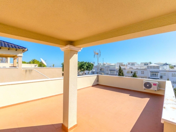 Sunlit rooftop terrace with terracotta tiles, beige walls and columns, overlooking white Mediterranean buildings under a bright blue sky, with an outdoor AC unit on the wall.