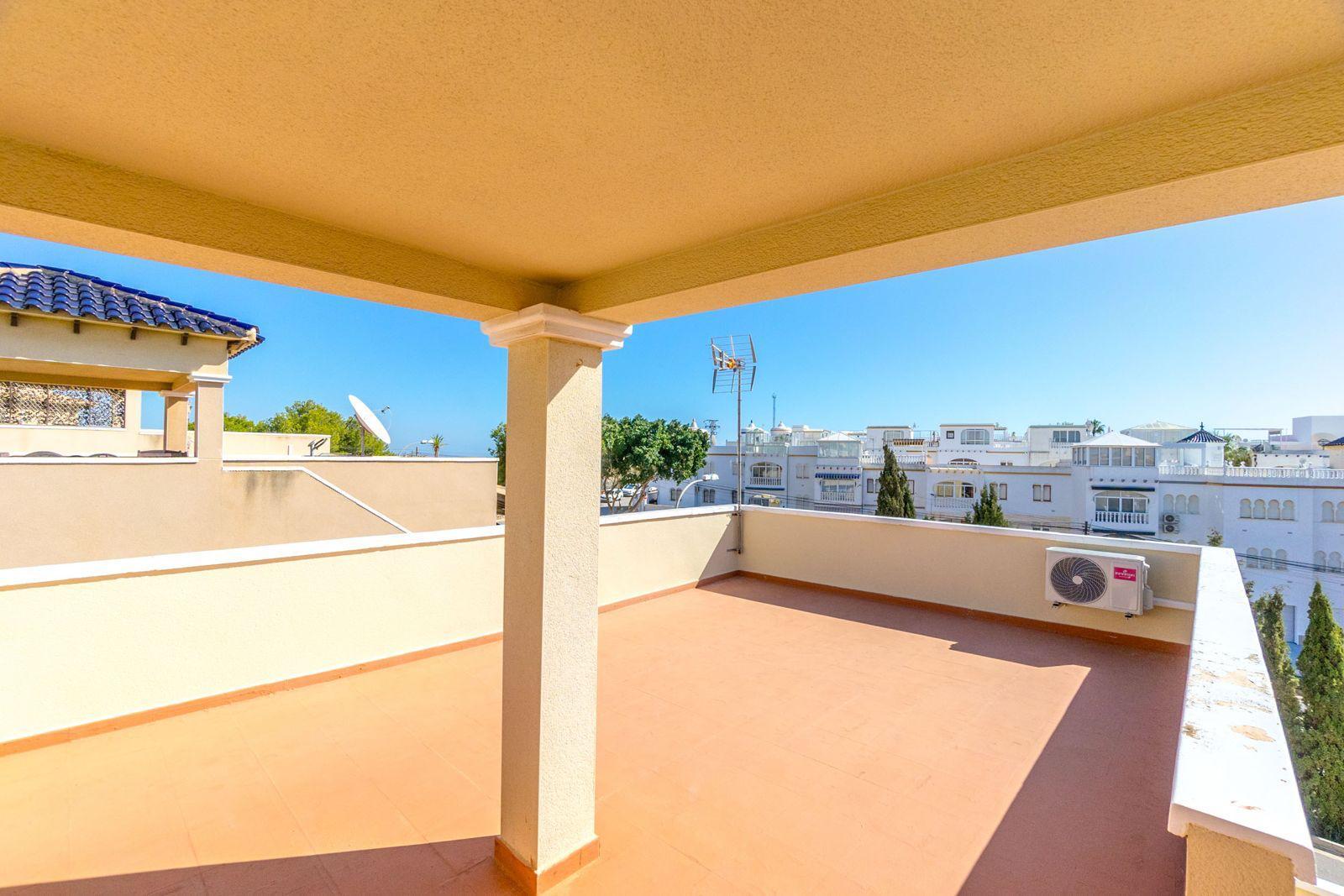 Sunlit rooftop terrace with terracotta tiles, beige walls and columns, overlooking white Mediterranean buildings under a bright blue sky, with an outdoor AC unit on the wall.