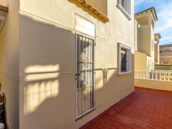 Beige exterior wall with a white security gate and barred window along a sunny red brick patio. A railing and neighboring buildings are visible in the background.