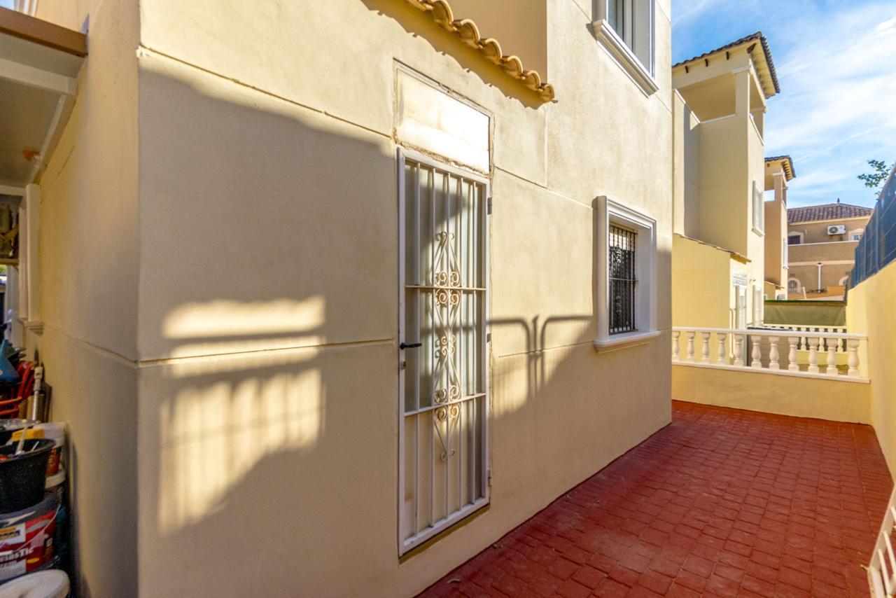 Beige exterior wall with a white security gate and barred window along a sunny red brick patio. A railing and neighboring buildings are visible in the background.