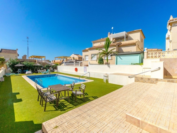 Poolside at a sunny resort: blue pool, wooden table with chairs on artificial grass, beige buildings in the background under a clear sky.