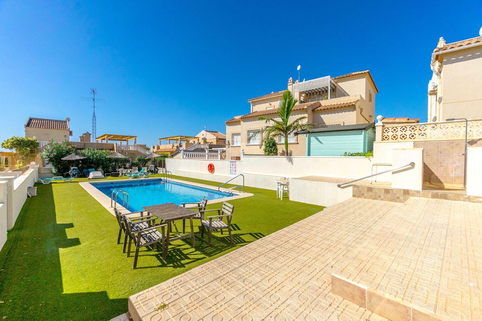 Poolside at a sunny resort: blue pool, wooden table with chairs on artificial grass, beige buildings in the background under a clear sky.