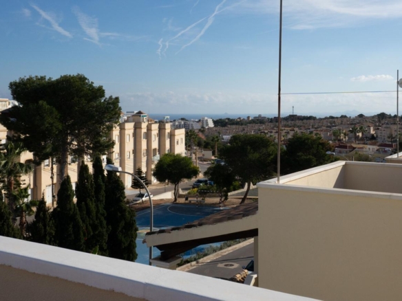 Balcony view over a sunny coastal town: beige apartment buildings, green trees, and a blue sky with the sea on the horizon.