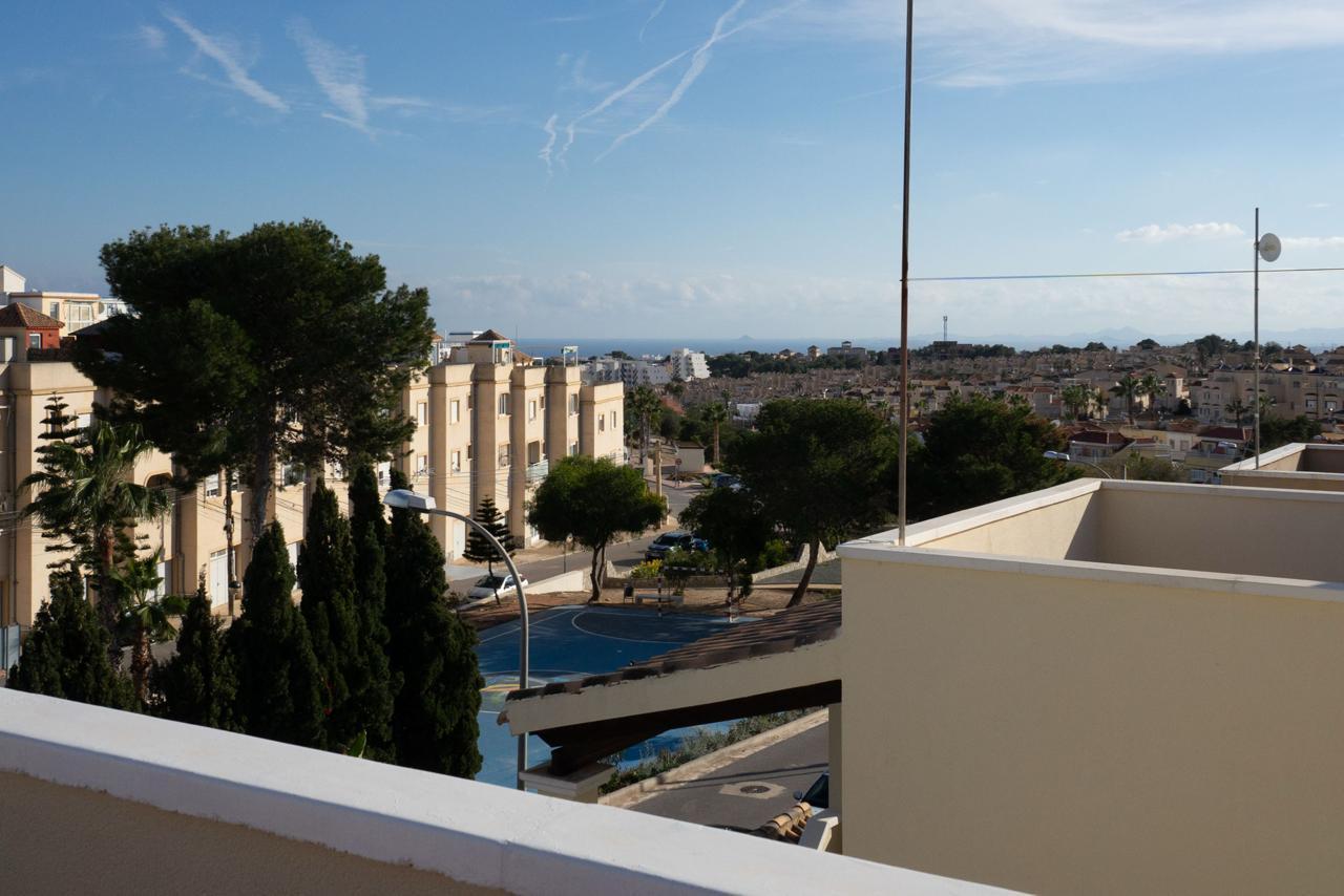 Balcony view over a sunny coastal town: beige apartment buildings, green trees, and a blue sky with the sea on the horizon.