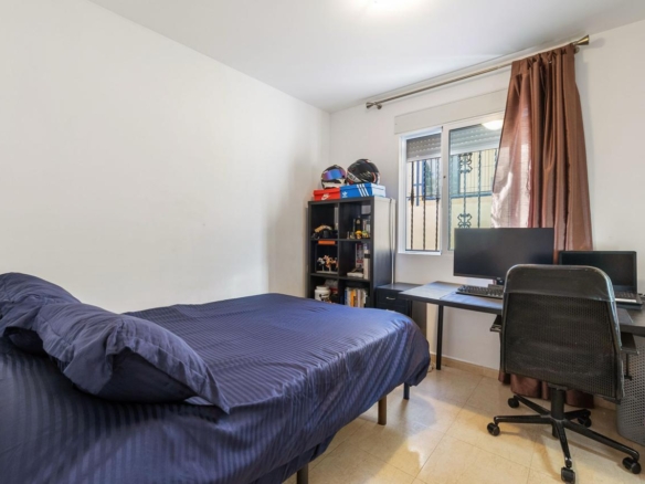 Small bedroom with a blue bed and pillows on the left, a black desk with dual monitors on the right, and a window with brown curtains in the corner.