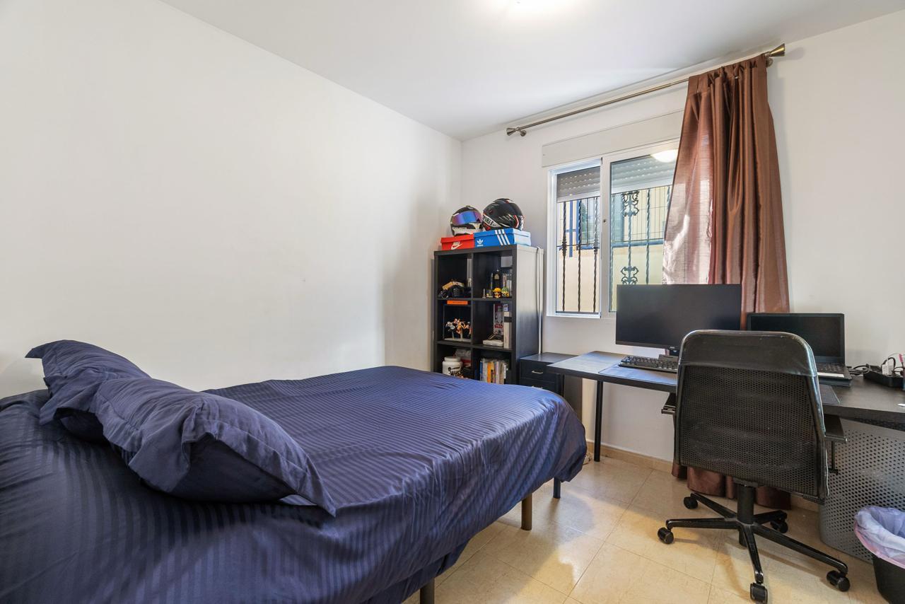 Small bedroom with a blue bed and pillows on the left, a black desk with dual monitors on the right, and a window with brown curtains in the corner.